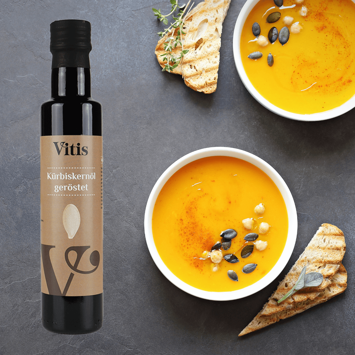 EIne Flasche Kürbiskernöl geröstet von Vitis neben zwei Schalen Kürbissuppe mit Toastbrot.