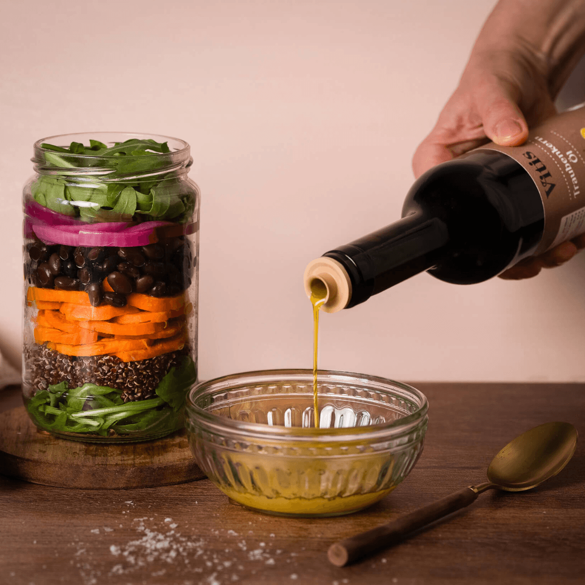 Vitis Traubenkernöl wird in eine Schale gegossen. Daneben ist ein Glas mit einem Schichtsalat zu sehen.