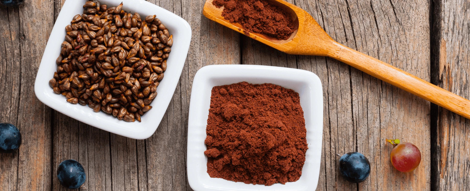 Zwei weiße Schalen dekorativ auf einem Holztisch angerichtet. In der einen Schale sind Traubenkerne und in der anderem Traubenkernmehl.