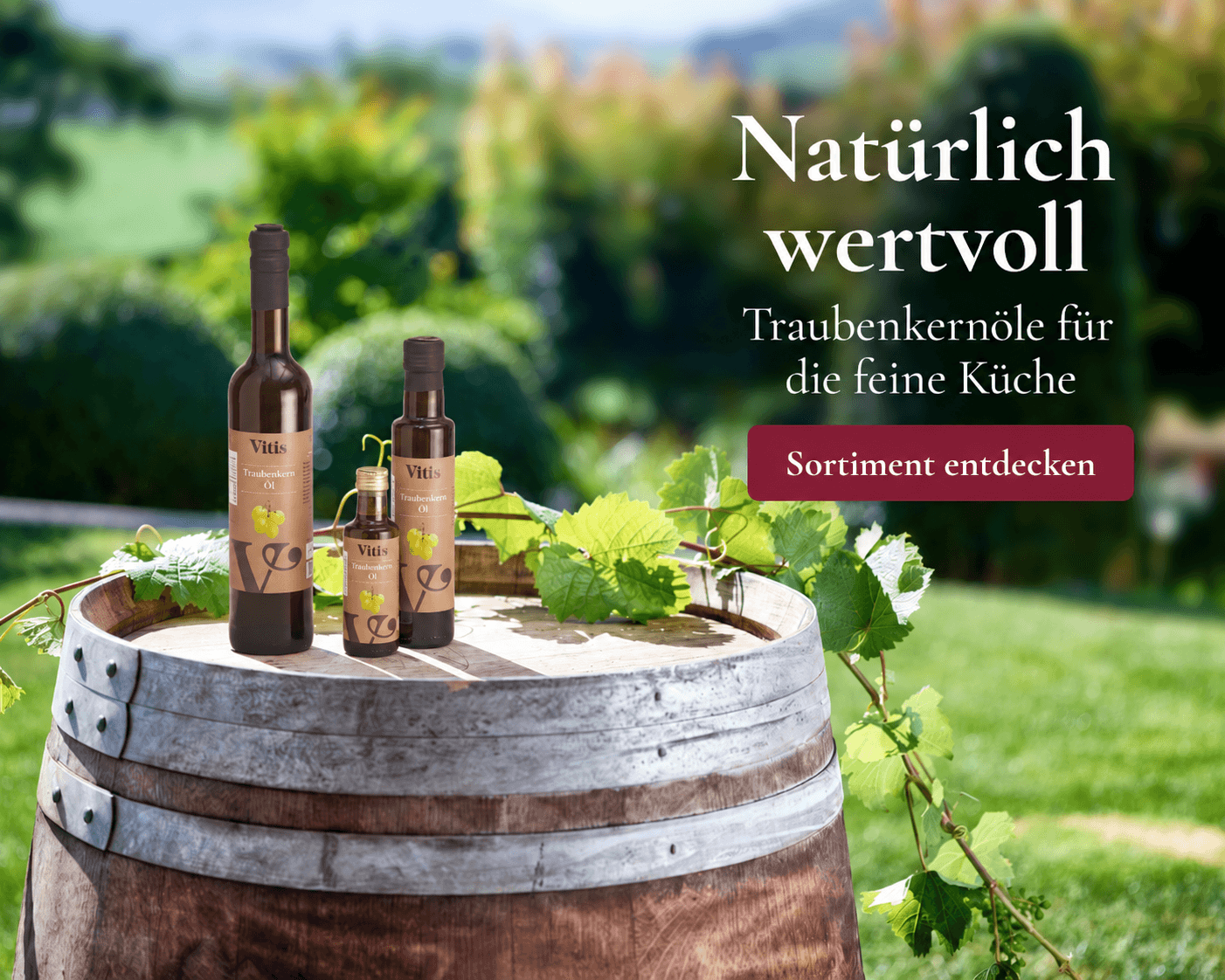 Vitis Traubenkernöle in drei Flaschengrößen auf einem Weinfass, dekoriert mit frischen Weinblättern.